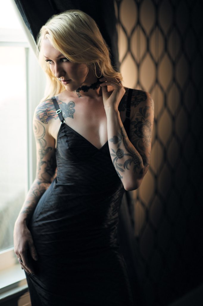 A tattoo boudoir photo from Sonoma County shows a blonde woman in a black slip dress with thin straps, standing sideways near a curtained window. Her pose involves one hand gently touching her lace choker, with intricate floral and animal tattoos visible on her arms. Soft natural light from the window highlights her hair and skin against the dark, patterned wall background, creating a moody composition in muted tones.