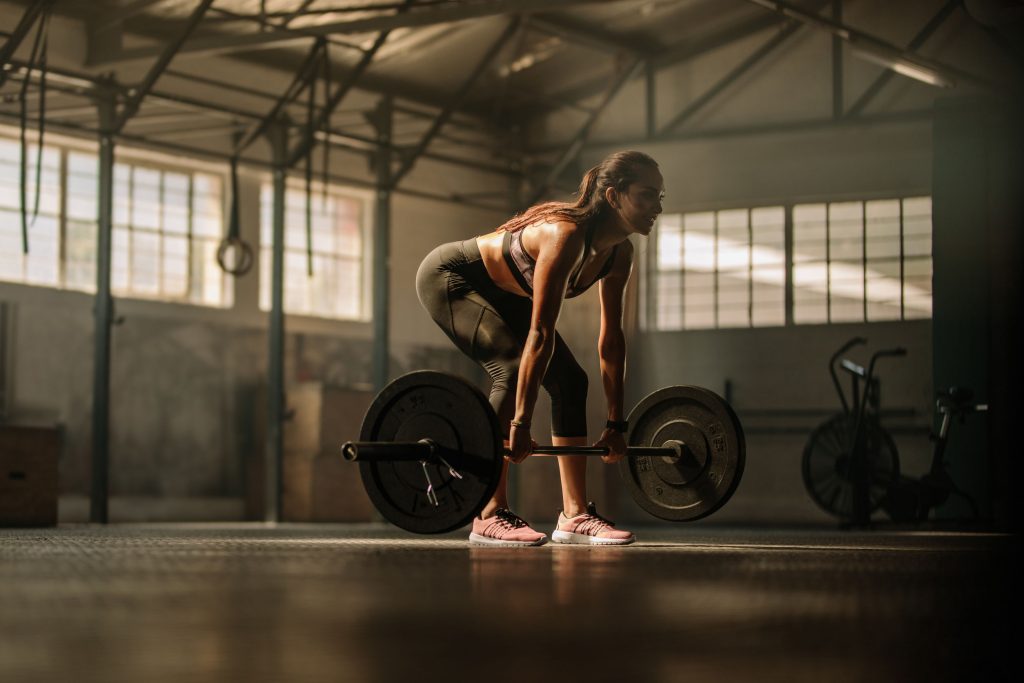 Image illustrating how to tone up for your boudoir photo shoot, with a woman in a bent-over pose lifting a weighted barbell in a spacious gym, natural sunlight from large windows creating warm golden highlights and soft shadows on neutral gray and brown tones; ponytail hairstyle, dressed in a pink sports top and dark leggings with pink sneakers, composition centering her focused stance amid blurred equipment like rings and bikes for an active atmosphere.