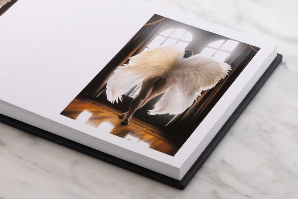 This angel wing boudoir album lays open on a white marble surface. The left page remains blank white while the right page displays a large printed photograph of a woman captured from behind in a graceful standing pose. She wears a voluminous white feathered angel wings that fan out dramatically around her lower body. Warm golden lighting illuminates the scene creating soft glowing highlights on the feathers and her skin while arched windows with intricate panes and rich brown drapery form an elegant grand interior background. 