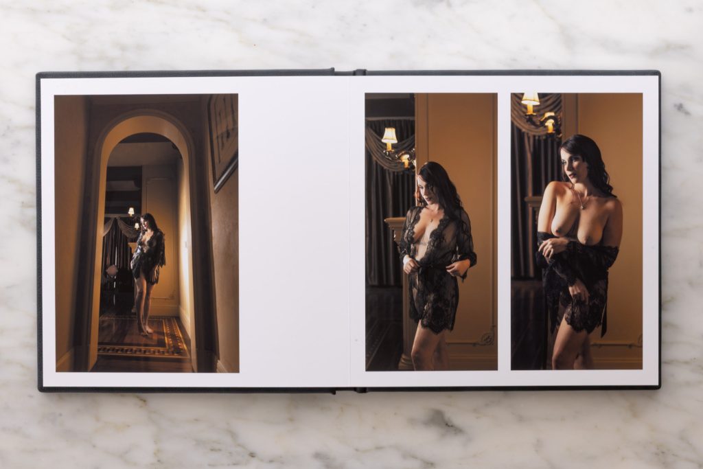 An artistic boudoir album design lies flat on a white marble surface displaying a multi-photo spread across both pages. The left page shows a vertical image of a woman standing barefoot in a long arched hallway with warm golden lighting casting soft shadows on ornate walls and patterned wooden floors while she wears a flowing black garment draped over one shoulder. The right page includes two vertical photographs of a woman with dark wavy hair posing gracefully in a luxurious interior with chandeliers and rich drapery. In the first she adjusts her sheer black lace attire with hands at her waist while soft amber light highlights her skin. In the second she crosses her arms modestly over her chest with the lace garment open revealing bare shoulders as warm tones create gentle contrasts throughout.