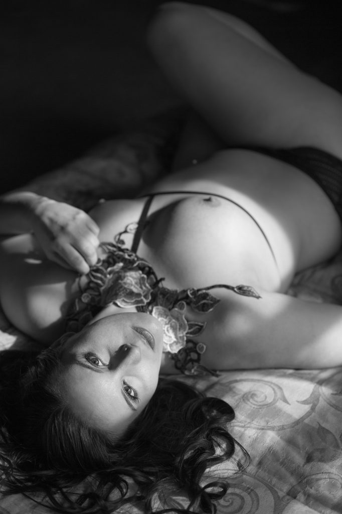 A woman with long dark hair reclines on a patterned bedspread during her artistic hard light nude portraits. Grayscale tones emphasize sharp contrasts and shadows illuminating her skin while she gently feels the floral neck and chest piece as the light falls on her face perfectly for a short light pattern in this serene pose amid soft fabric folds within the intimate composition.