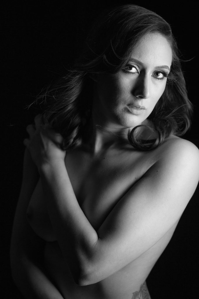 A woman with long wavy hair gazes upward during her artistic implied nude photography session. One arm crosses over her waist while the other covers her chest. Dramatic black and white lighting creates deep contrasts and soft shadows on her bare skin against a solid dark background.