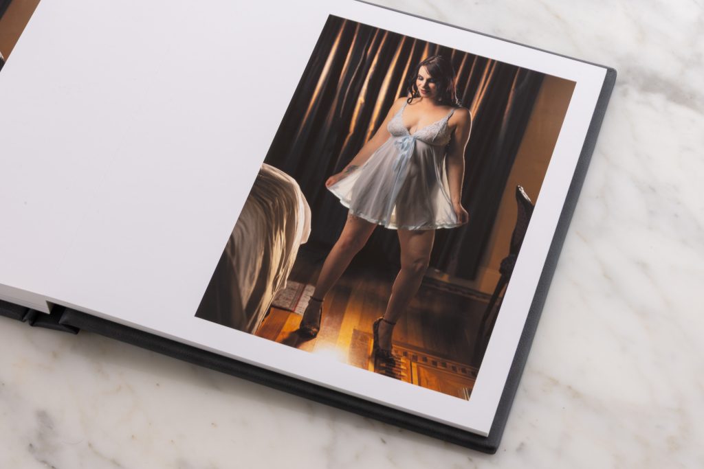 An artistic lingerie album spread showing an image on the right page of A woman with dark wavy hair standing playfully with arms extended holding the skirt end of her light blue sheer babydoll nightie. Warm golden lighting casts soft amber glows and subtle shadows across her skin and the bedroom setting with dark curtains, a bed, and polished wooden floors while she balances on strappy high heels. 