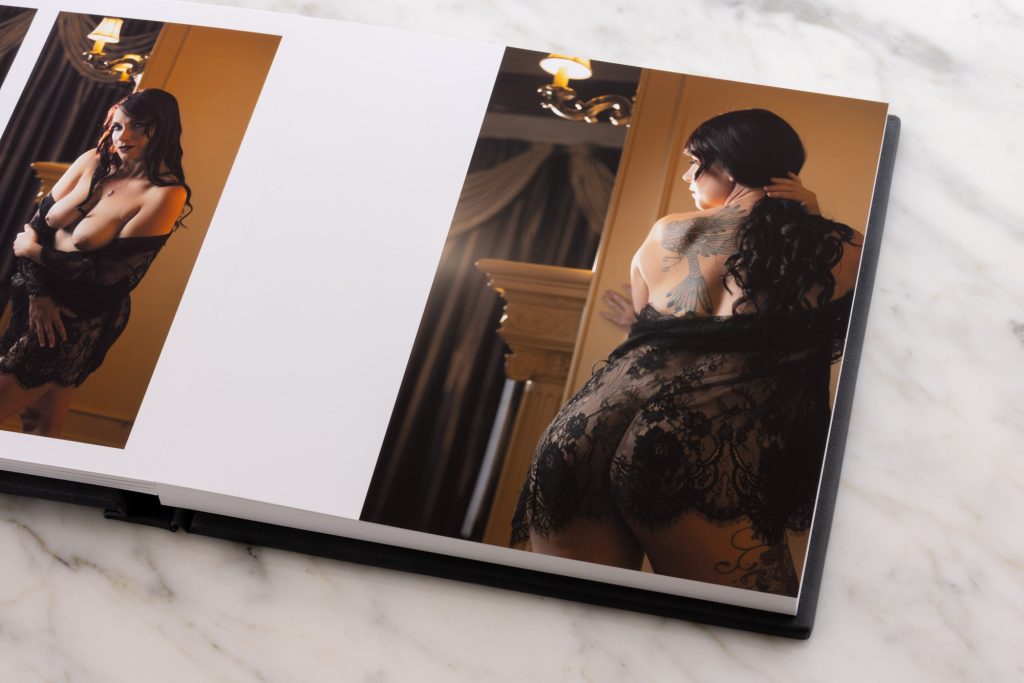 An artistic tattoo boudoir albums displays two vertical photographs on the left and right pages while resting on a white marble surface. The left image shows a woman with long dark hair standing gracefully with arms crossed over her chest as a sheer black lace robe drapes loosely from her shoulders. Warm golden chandelier lighting creates soft amber glows and subtle shadows across her skin and the elegant curtained interior. The right photograph captures a woman viewed from behind with one hand gently brushing her hair to the side to reveal a detailed wing tattoo spanning her upper back. She wears delicate black lace attire that complements the intricate ink while soft yellow tones illuminate the ornate room details.
