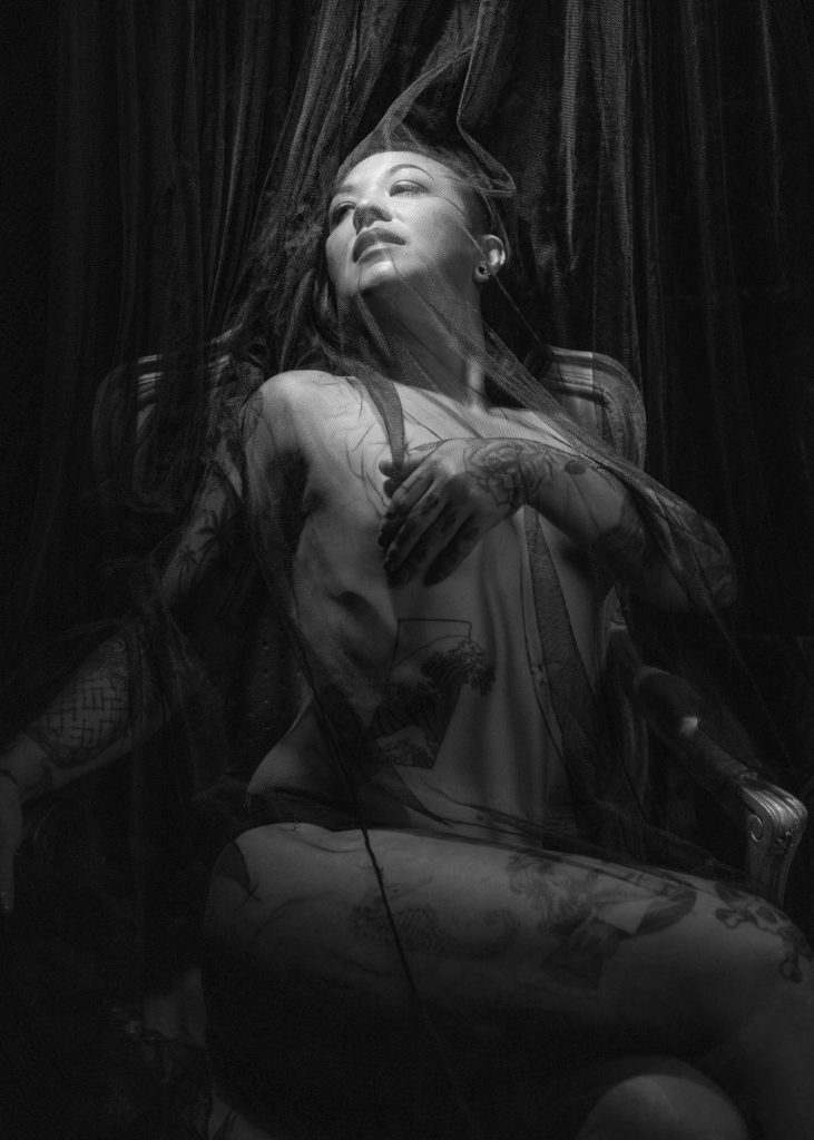 A woman with long dark hair reclines gracefully behind a sheer black veil during artistic tattoo photo session. She places her hand on her chest in a serene upward gaze pose, intricate tattoos covering her arms and torso visible through the translucent black tulle fabric, while the black-and-white composition features dramatic contrast with concentrated light on her face and fast fall off into shadow.