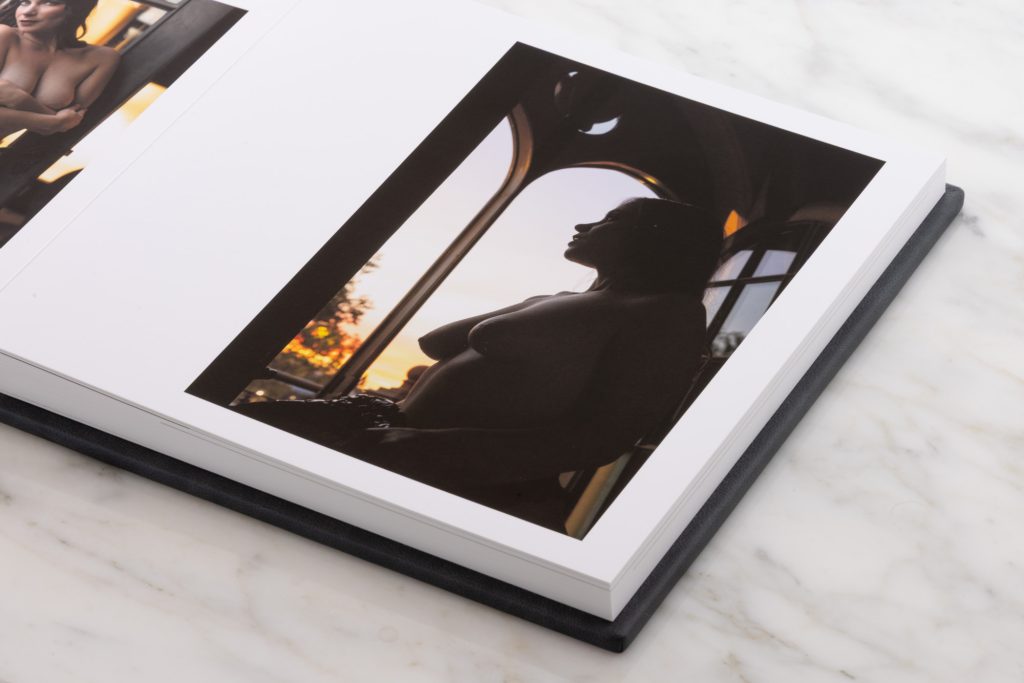 An open boudoir album with silhouette image on the right page rests on a white marble surface. The left page shows a small color photograph of a woman in profile with arms crossed over her chest and bare shoulders illuminated by warm golden light from windows. The right vertical image captures a dark silhouette of a woman gazing upward toward an arched window at sunset creating dramatic contrasts with orange and yellow hues glowing through the glass panes and reflecting on the floor. Thick white pages and indigo blue leather binding frame the compositions elegantly highlighting the soft backlighting and rich warm tones throughout.