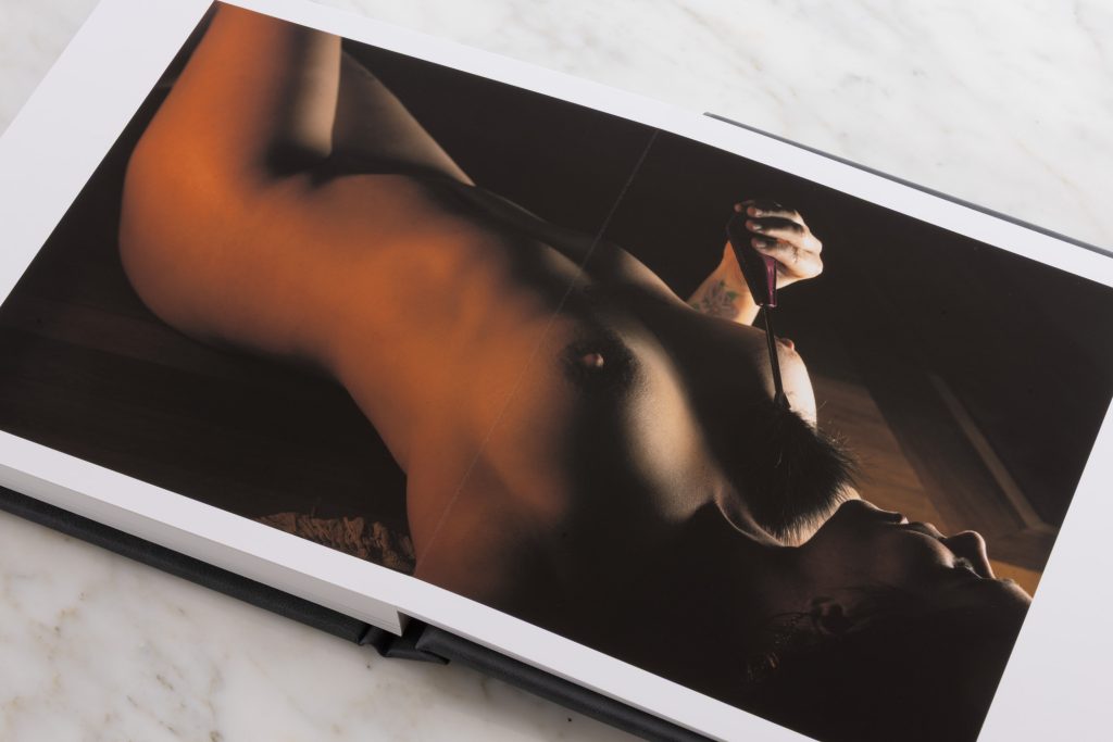 A dramatic shadow artistic boudoir album displays a horizontal photograph across both pages while resting on a white marble surface. The image shows a woman reclined on her back with one arm extended holding a small feather. Warm amber lighting casts intense contrasts and golden highlights on her bare skin revealing a colorful tattoo on her arm while the dark background emphasizes deep brown tones and subtle contours. 