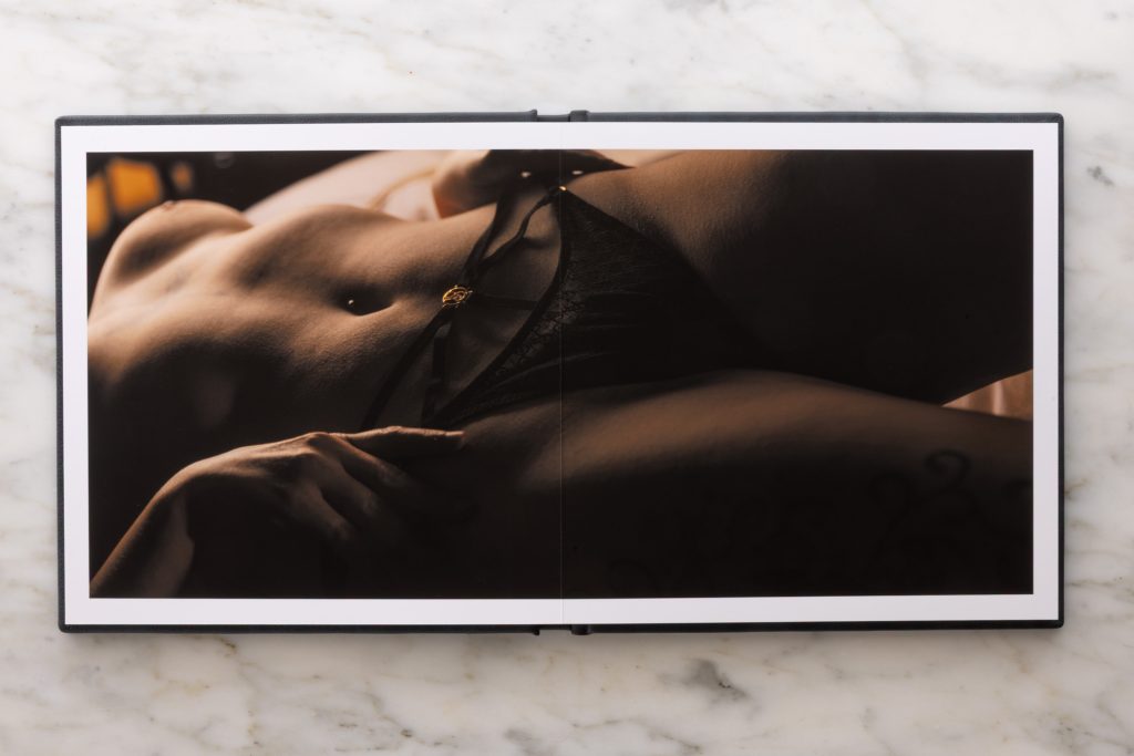 a horizontal photograph extending across both pages in this fine art boudoir album spread while resting on a white marble surface. The image captures a close-up view of a woman's reclined torso with one hand gently placed near her hip. She wears intricate black lace bottoms accented by a small gold Victoria's Secret emblem at the center. Dramatic warm lighting in deep amber and brown tones creates soft shadows and glowing highlights on her skin revealing a subtle swirling tattoo on her side. 