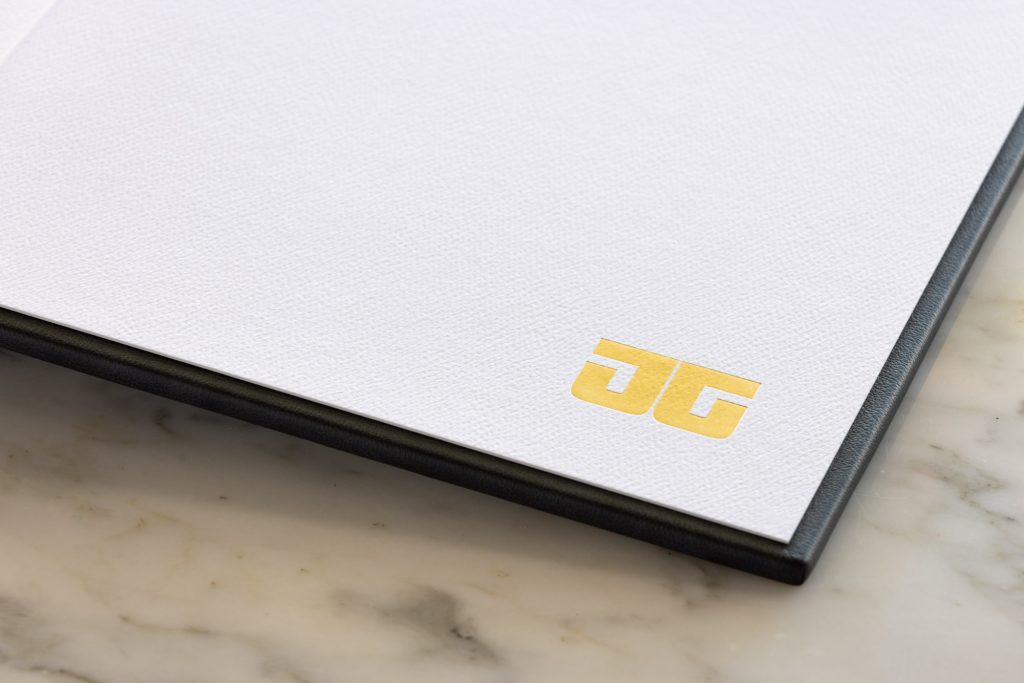 A close-up view shows the end papers of a premium boudoir album. The white textured pages feature a striking gold foil JG logo positioned in the lower right corner of the last page. Indigo blue leather edges frame the clean minimalist design. 