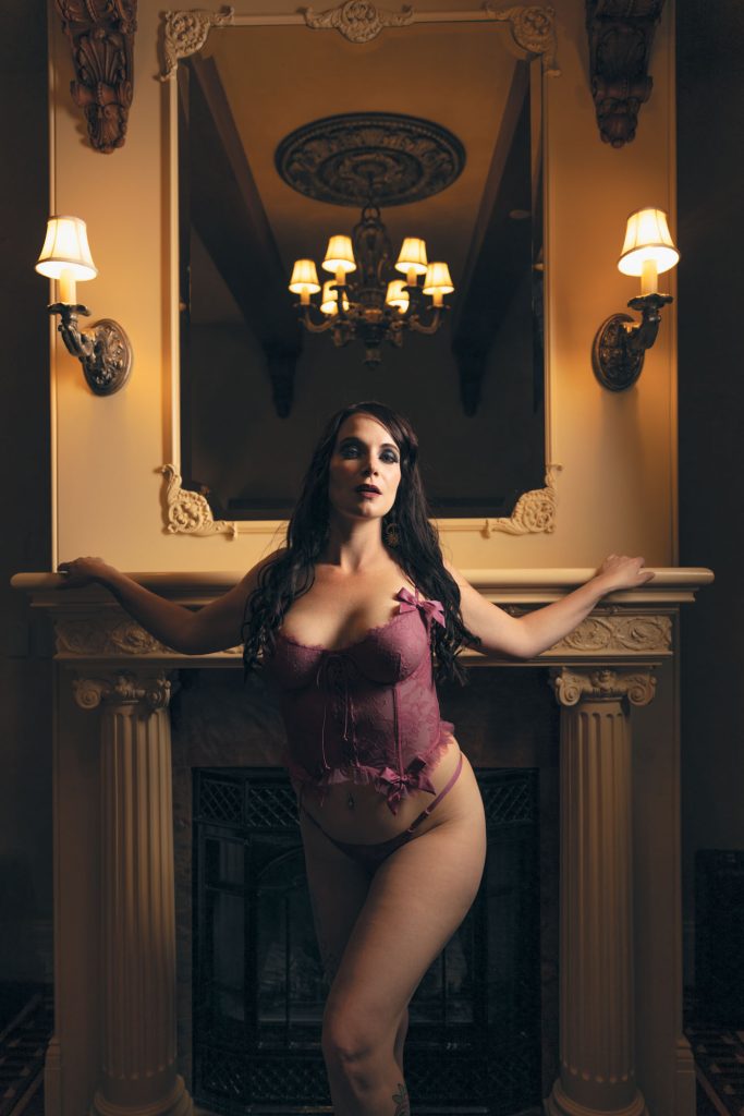 A woman with long dark hair poses for her gothic boudoir photos in Sonoma County while standing before an ornate fireplace with arms extended along the mantel. She gazes directly forward in a balanced stance illuminated by warm golden lights from wall sconces and a chandelier reflected in the large mirror above. Deep maroon lace attire complements the elegant cream tones of the carved columns and detailed room accents.