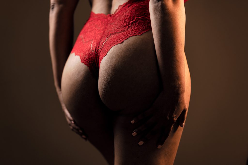 Example showing how to take tasteful booty pics featuring an Indian woman from behind. She stands with both hands placed gently on her lower thighs while her body angles slightly away from the camera. Warm dramatic lighting creates soft highlights and shadows across the composition against a neutral background.