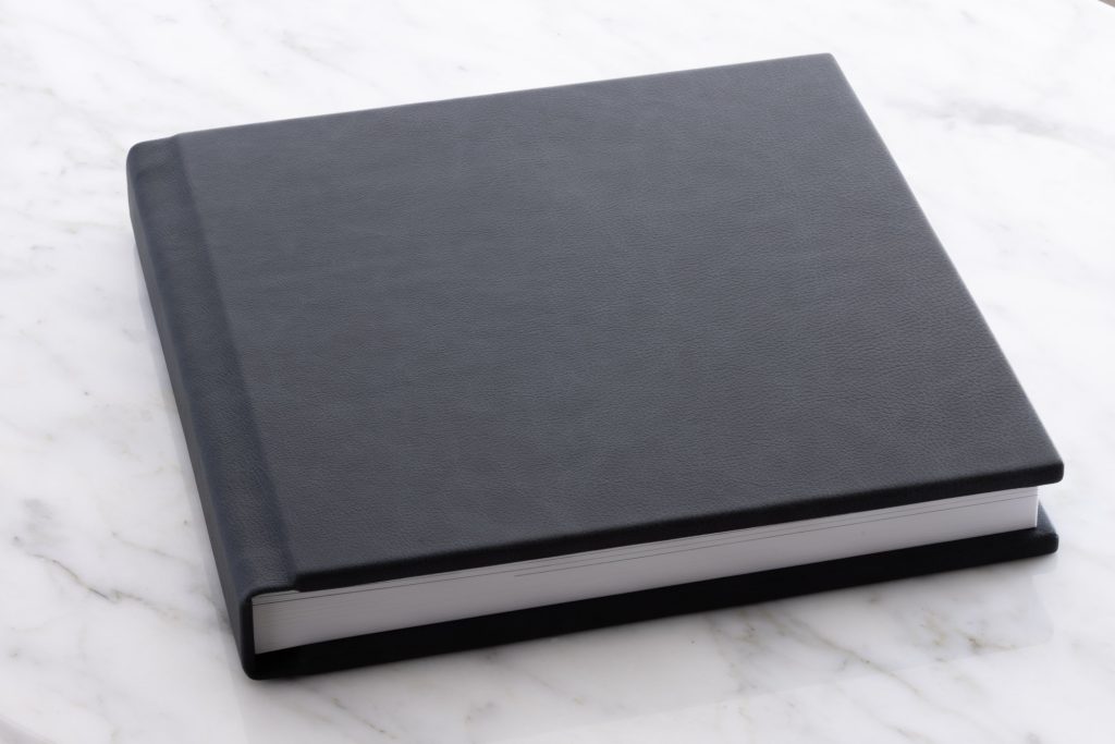A closed indigo blue leather-bound boudoir album resting on a white marble surface. The album features a smooth textured cover in deep charcoal with subtle grain visible in the leather. Its square format includes clean straight edges, a crisp white interior page edge along the bottom, and a simple elegant design with no visible text or embellishments on the front. 