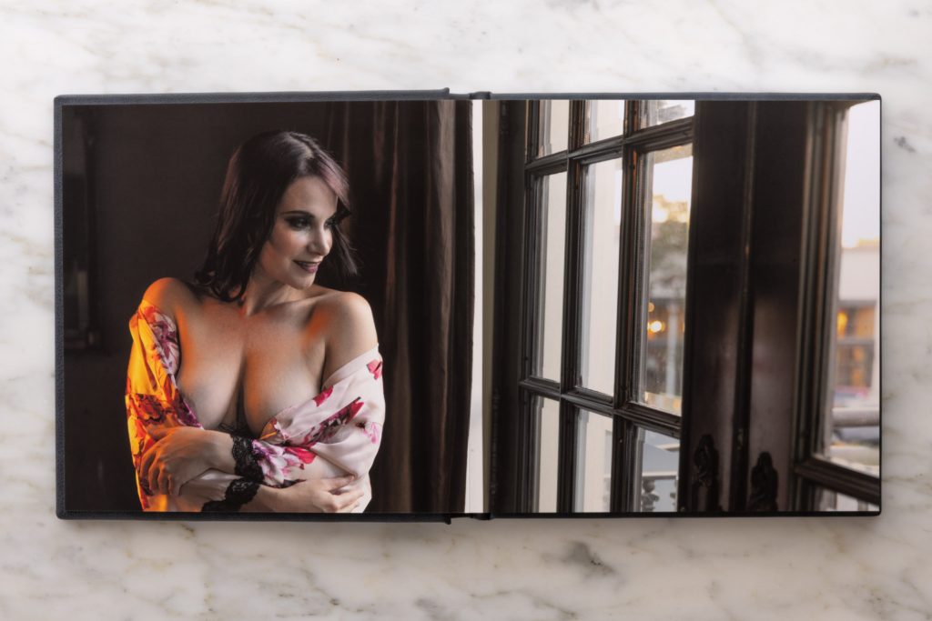 An open layflat boudoir album lies flat on a white marble surface displaying a full double-page spread photograph. A woman with dark hair and striking eye makeup gazes slightly to the side with a gentle smile. She wears a floral-patterned robe in soft peach and pink tones that drapes loosely off her shoulders revealing bare arms crossed modestly over her chest.