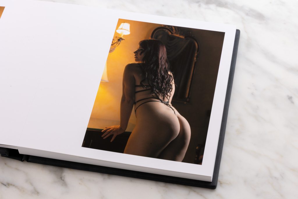 A luxury boudoir photo book lays open showcasing a single vertical photograph on the right page while placed on a white marble surface. A woman with long wavy dark hair appears in profile from behind as she leans gently forward with one hand resting on a desk. She wears slim black straps that cross her back. Soft warm yellow lighting from a wall sconce casts golden highlights and subtle shadows across her skin and the ornate mirror in the dim elegant interior background. 