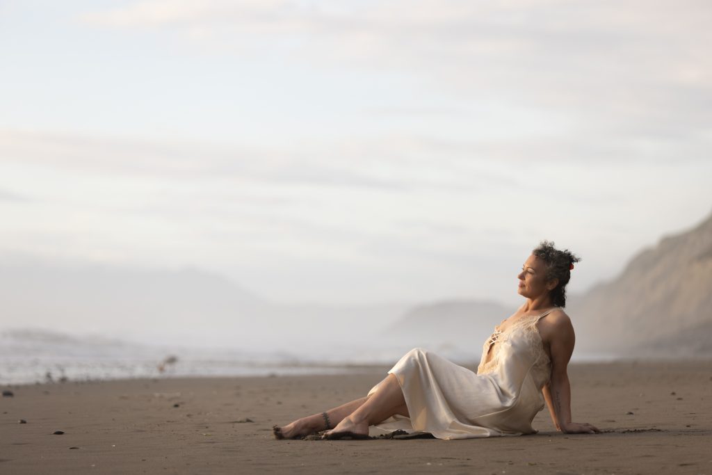 A woman with curly dark hair poses for her outdoor lingerie photo shoot on a sandy beach. She reclines leaning on one arm with her bare legs extended as she gazes toward the ocean. She wears a flowing cream colored satin dress with delicate lace details. Soft diffused lighting creates a serene atmosphere with misty mountains and ocean waves visible in the background.