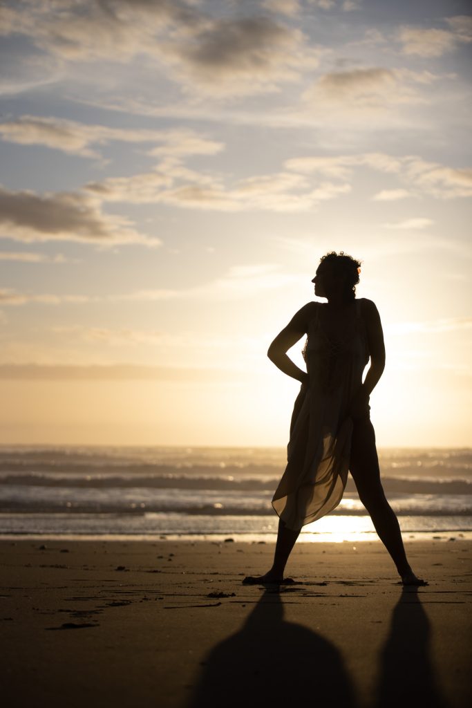 A woman with curly dark hair poses for her outdoor silhouette boudoir photos at sunset on a sandy beach. She stands barefoot with legs apart and one hand on her hip while looking out toward the ocean. Warm golden light from the setting sun creates a striking silhouette of her in a flowing dress against the dramatic sky and waves with a long shadow cast on the sand.