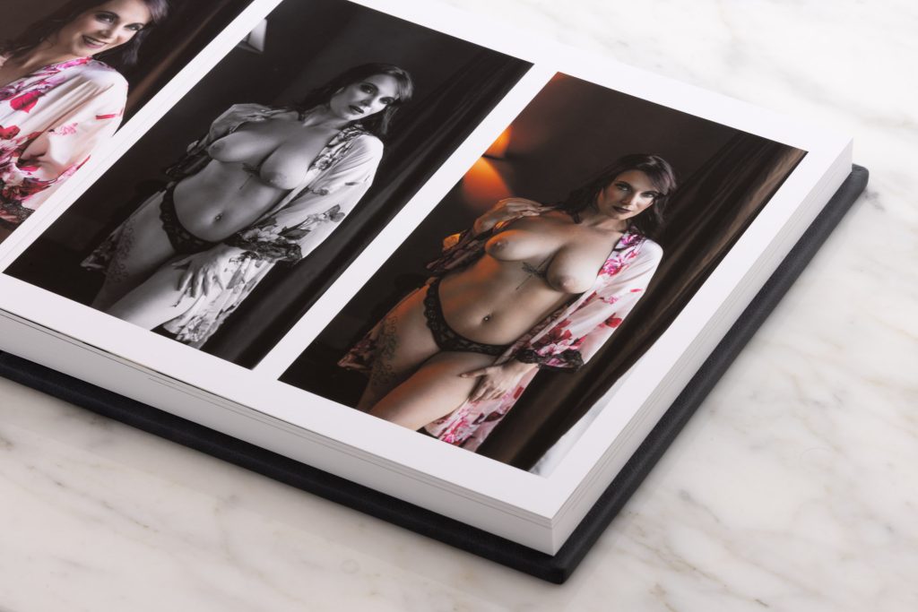 An open album reveals triptych boudoir album design featuring three vertical photographs arranged side by side across a two page spread. The left color image presents a woman with dark hair smiling softly while wrapped in a vibrant floral robe in pink and orange hues. The center black-and-white photograph shows her in a thoughtful side profile with the robe draped loosely over her shoulders and black lace bottoms. The right color photo captures her gazing directly forward with one hand on her hip and the floral robe open revealing intricate tattoos and a navel piercing.