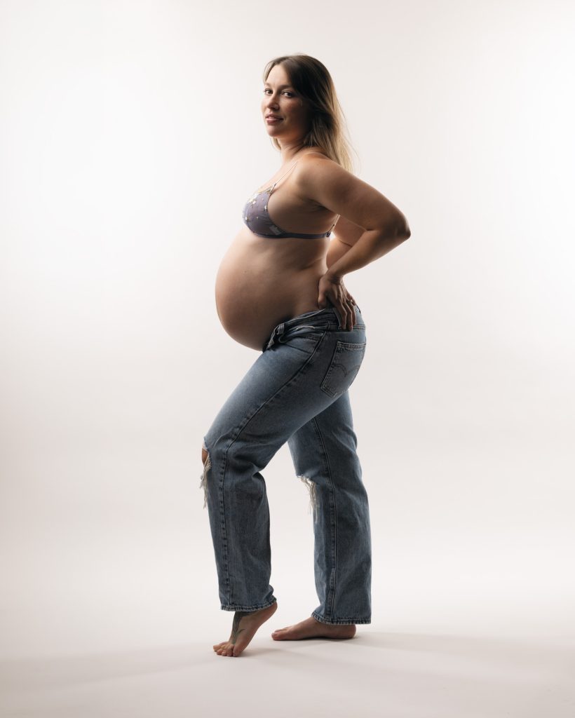 A pregnant woman stands confidently in a bright studio setting for her bay area maternity photos. She turns her body slightly while looking back over her shoulder toward the camera with a gentle smile. One hand rests on her hip as she highlights her rounded belly in a relaxed pose. Soft even lighting illuminates the natural colors of her light purple floral lace bra and blue ripped denim jeans. She remains barefoot against the seamless white backdrop.