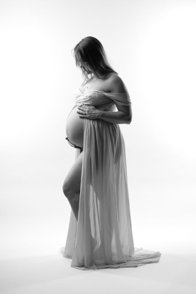 A pregnant woman stands in a bright studio setting for her black and white maternity photos. She cradles her rounded belly gently with both hands while gazing downward in a tender pose. Dramatic lighting creates soft highlights and shadows across the image emphasizing the flowing sheer fabric of her dress. The open front design drapes elegantly around her form with the long train pooling on the floor. She remains barefoot against the seamless white backdrop.