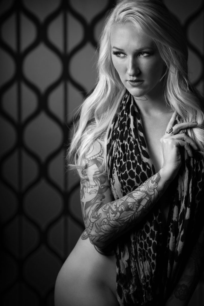 Boudoir hairstyles inspiration featuring a woman with long blonde hair and extensive tattoos. She poses for her session as she gazes directly at the camera with one hand touching her hair while draped in a leopard print garment against a geometric patterned background in dramatic black and white lighting.
