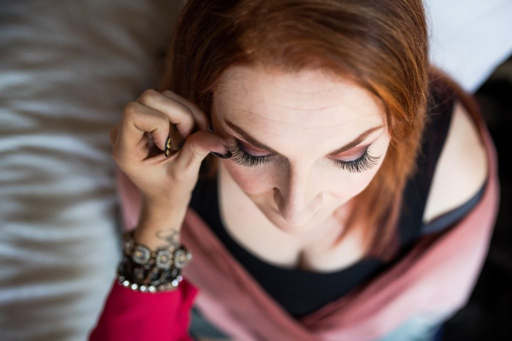 Makeup artist helps a woman with red hair carefully apply a woman's eyelashes during her boudoir makeover on white bedding. Her head is tilted downward as she uses her hand with black nail polish to finesse the lash near her eye which has long lashes and pink eyeshadow the woman wears a red top layered over black with a pink scarf while soft lighting highlights the details of her makeup and accessories in the tight composition.