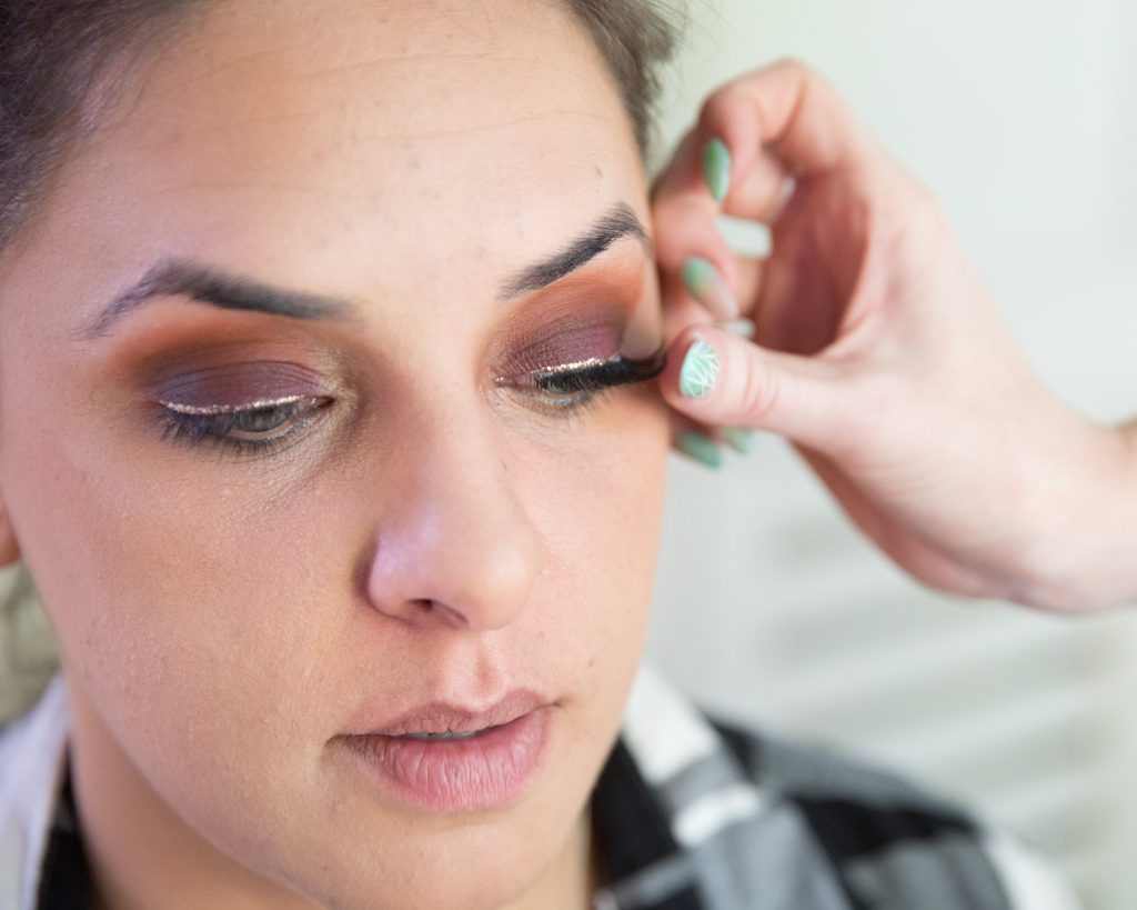 A makeup artist carefully applies a set of false strip lashes during a boudoir strip lash application. The woman’s eyelids are adorned with warm, smoky eyeshadow in rich copper and deep brown tones, blended seamlessly across the crease. A thin line of metallic silver eyeliner traces her upper lash line, adding a touch of shimmer to the bold eye look. The makeup artist’s hand, with nails painted mint green and featuring delicate white line art, gently presses the strip lash into place. The dark, voluminous lashes instantly enhance the woman's eyes, complementing the sultry boudoir makeup. Her lips are bare with a natural pink hue, and her complexion has a soft matte finish. The close-up captures the precision and artistry involved in the lash application, highlighting the transformation of the eyes into a focal point of the boudoir look.
