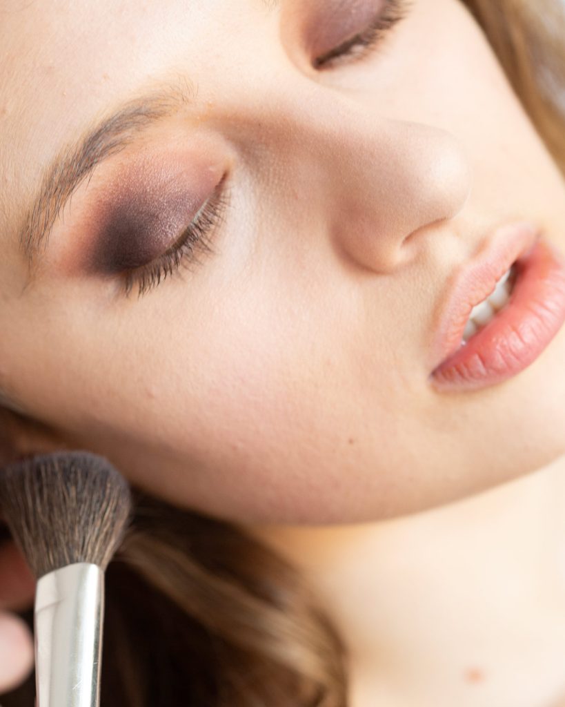 A close-up captures the delicate process of boudoir makeup foundation application. A soft, fluffy makeup brush gently sweeps across the woman’s cheek, blending foundation to create a smooth, even complexion. Her skin appears radiant and natural, with a subtle glow highlighting her cheekbones. Her eyes are adorned with a smoky eyeshadow in shades of deep brown and soft mauve, seamlessly blended to create depth and drama. Her eyelashes rest softly against her skin as her eyes remain closed during the application. Her lips are a soft, natural pink, adding to the fresh and effortless look. Her hair cascades in soft waves, partially visible in the background. The lighting is bright and natural, emphasizing the flawless finish achieved through the boudoir makeup foundation application.