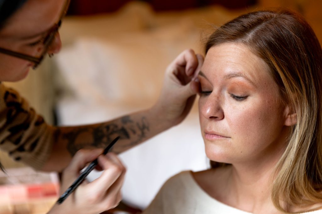 In this intimate preparation scene, a woman with light skin and straight, blonde-highlighted brown hair sits serenely with her eyes closed as a makeup artist applies eyeshadow to her upper eyelids. The woman wears an off-the-shoulder, cream-colored sweater that subtly reveals her collarbone, contributing to the soft, sensual tone of the moment. Her complexion is even and naturally radiant, enhanced by minimal foundation and a touch of blush. The makeup artist, visible in the foreground with a tattooed arm and wearing glasses, uses a fine brush to blend a warm, copper-toned shadow onto the eyelid, creating depth while maintaining a soft, romantic finish. The gentle lighting and neutral-toned background, possibly a bed with white sheets and a beige headboard, add to the relaxed, cozy atmosphere. This application scene perfectly illustrates Boudoir makeup tips for better photos, emphasizing the importance of warm, flattering tones and professional blending to enhance natural features without overpowering them.