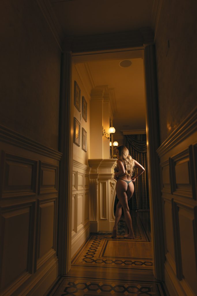 A woman with long blonde hair poses for her bay area boudoir photography session while standing in an elegant hallway with her back to the camera. She places both hands on her hips and wears a dark teal lace bra and matching bottoms. Warm golden light from wall sconces illuminates the dark wood paneling and geometric patterned floor that leads toward an ornate fireplace visible in the background.