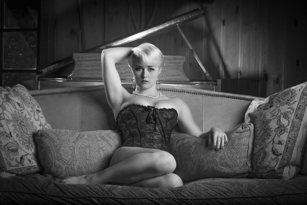 A woman with short blonde hair performs boudoir poses on couch while seated on a large patterned sofa in this black and white photograph. She wears a lace corset top paired with a pearl necklace and earrings with one arm raised behind her head and her other hand resting on a pillow as she looks directly at the camera. A grand piano is visible in the wood paneled background.