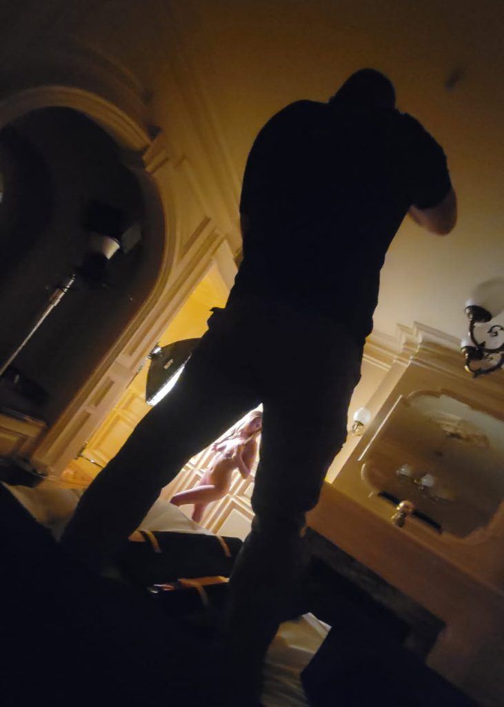 A photographer in black clothing stands with legs apart in the foreground while capturing a woman during her boudoir session. She poses against an intricate wall in the background with blonde hair visible through an arched opening as warm golden lighting casts soft shadows across the luxurious room featuring a fireplace mantel and mirror in the low angle composition.
