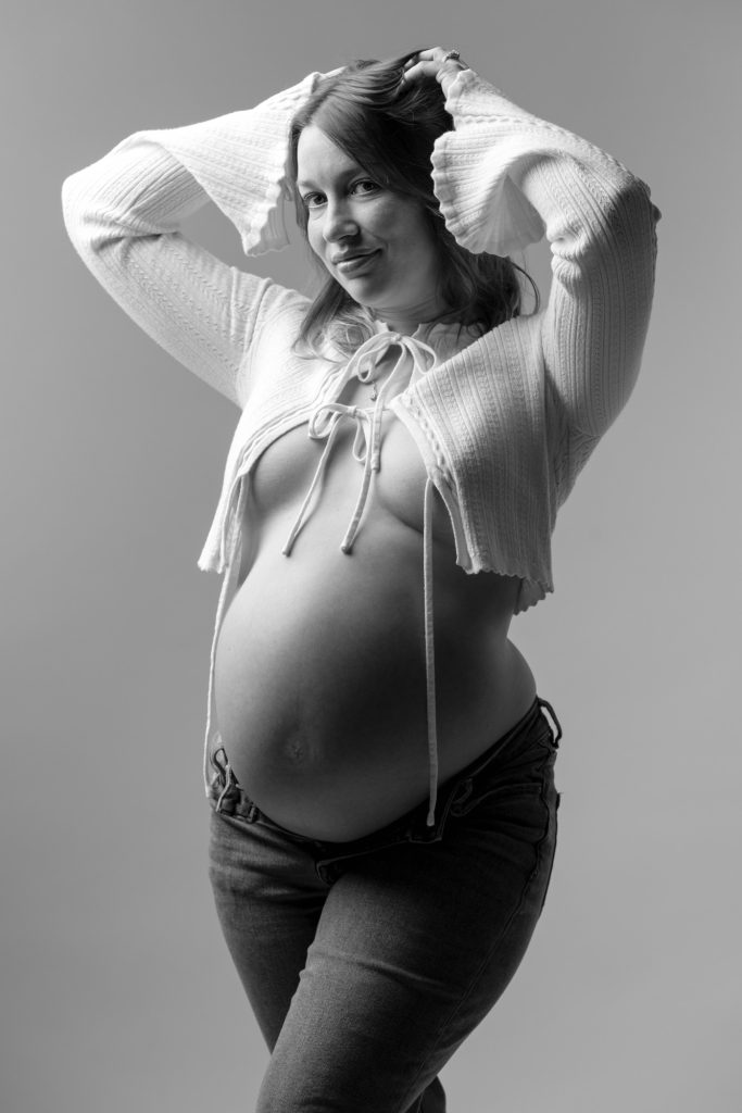 A pregnant woman poses confidently in a black and white studio setting for her Calvin Klein maternity photos. She stands with both hands raised to her hair while looking toward the camera with a gentle smile. Dramatic lighting creates strong highlights and shadows across the image emphasizing her rounded belly. She wears an open white knit cardigan with tie details paired with dark jeans.