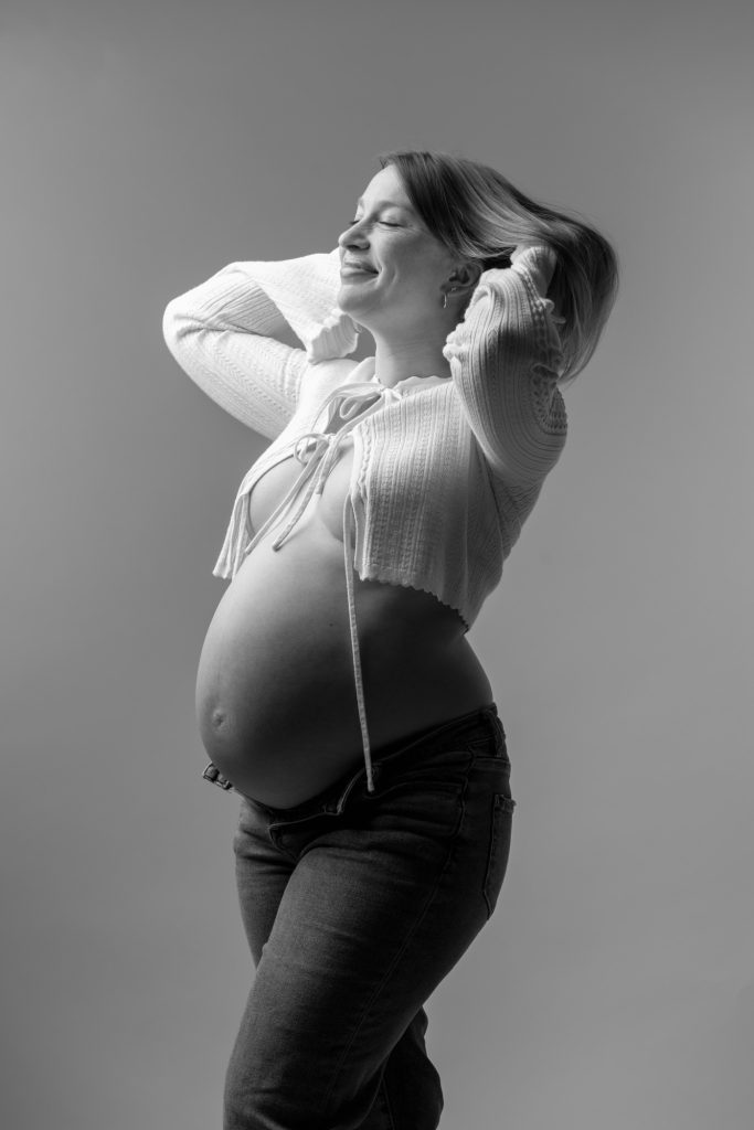 A pregnant woman poses joyfully in a black and white studio setting for her casual maternity photos. She stands with both hands raised to her hair while tilting her head back with eyes closed and a warm smile. Dramatic lighting creates strong highlights and shadows across the image emphasizing her rounded belly. She wears an open white knit cardigan with tie details paired with dark jeans.
