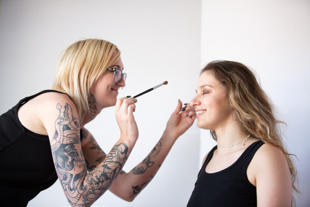 A tattooed makeup artist with short blonde hair, glasses, and a septum piercing applies eyeshadow to a smiling client in a minimalist, well-lit space. The artist, wearing a black sleeveless top, leans in with a focused yet friendly expression, holding a blending brush near the client’s closed eyelid. Her arms feature bold black tattoos, including a detailed portrait of a woman with tears and a coiling snake. The client, with wavy, light brown hair, wears a black tank top and a delicate silver necklace, appearing relaxed and happy. The scene captures the intimate, artistic process of client eyeshadow application, emphasizing skill, trust, and creativity in makeup artistry.