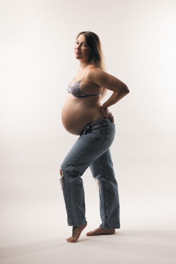 A pregnant woman stands in a bright studio setting during her cute maternity boudoir session. She turns her body slightly while looking back over her shoulder toward the camera with a gentle smile. One hand rests on her hip and the other supports her rounded belly in a relaxed pose. Soft even lighting illuminates the natural colors of her light purple floral lace bra and blue ripped denim jeans. She remains barefoot against the seamless white backdrop.