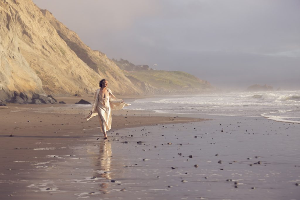 A woman with dark curly hair walks down the coast for her Daly City beach boudoir session while walking along the wet sand of a misty shoreline. She holds open a sheer robe over her flowing cream dress and smiles as waves crash nearby with dramatic cliffs rising in the background. Soft natural light creates gentle reflections on the sand and a serene mood along the ocean edge.