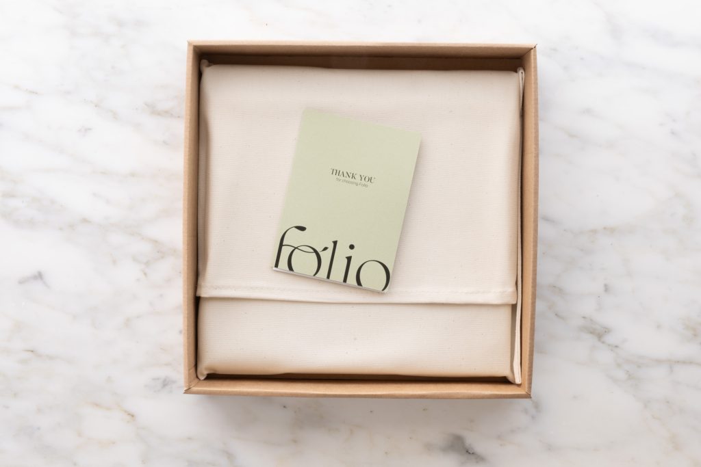 This is what you can expect when your boudoir photos are delivered. The photo shows a brown cardboard box on a marble surface showcasing a cream fabric pouch topped with a light green thank you card. The card features the word folio in stylized text while bright lighting illuminates the neutral colors and centered arrangement in the top down view.