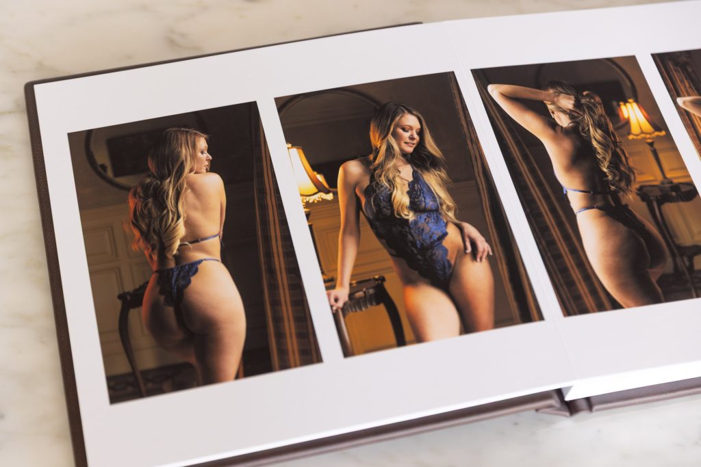 An open boudoir album rests on a marble surface showing the results of designing your boudoir album after a JG Boudoir session. Three photographs span across two pages of a woman with long blonde hair wearing blue lace lingerie in different poses and elegant room settings with warm lighting illuminating the white pages and brown cover in the composition.