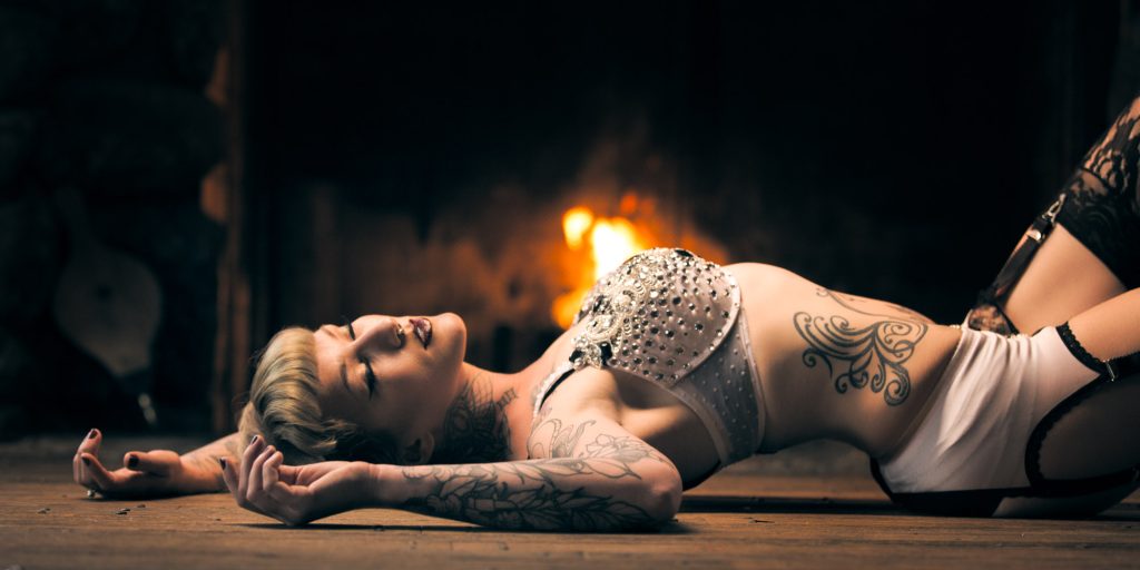 Model Ducky Rickman poses for some boudoir makeup inspiration. In this photo she has short blonde hair and extensive tattoos. She lies on her back on a wooden floor in front of a glowing fireplace dressed in a sparkling embellished white top and white bottoms with black lace accents. Her arms extend gracefully beside her as warm firelight dramatically illuminates the dark room.