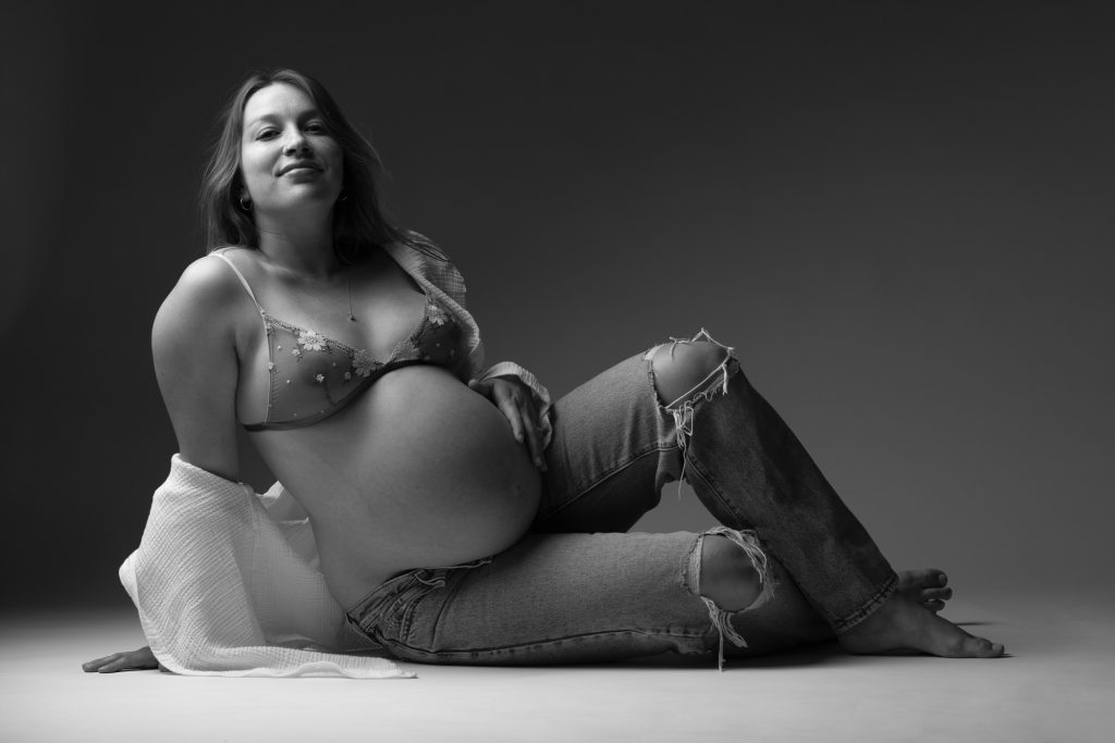 A pregnant woman reclines on the studio floor during her editorial style maternity photography session. She leans back supported on one arm with her other hand gently resting on her rounded belly while smiling softly toward the camera. Dramatic side lighting creates deep shadows and highlights across the black and white composition emphasizing her pose and curves. She wears a delicate lace bra top paired with ripped denim jeans and has a white textured wrap draped beside her while remaining barefoot against the minimalist gradient backdrop.