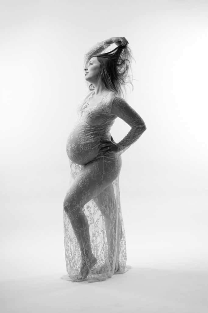 A pregnant woman stands gracefully in a bright studio setting for her lace dress maternity boudoir photos. She poses in profile with one hand on her hip and the other raised to lift her flowing hair while gazing upward in a confident expression. Dramatic lighting creates strong highlights and shadows across the black and white image emphasizing the intricate lace patterns of her long fitted dress. The sheer fabric drapes elegantly around her rounded belly. She remains barefoot against the seamless white backdrop.