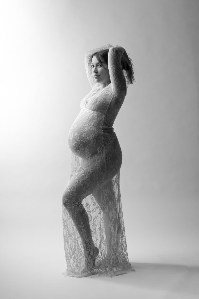 A pregnant woman poses gracefully in a bright studio setting during her maternity boudoir studio session. She stands with one hand raised to lift her flowing hair while looking toward the camera in a confident expression. Dramatic lighting creates strong highlights and shadows across the black and white image emphasizing the intricate lace patterns of her long fitted dress. The sheer fabric drapes elegantly around her rounded belly. She remains barefoot against the seamless white backdrop.