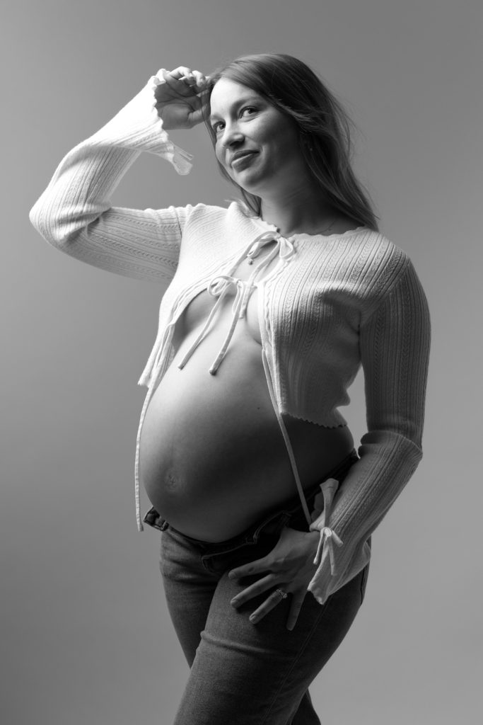 A pregnant woman poses confidently in a black and white studio setting for her maternity photos in jeans. She stands with one hand raised to her hair and the other resting near her hip while smiling warmly toward the camera. Dramatic lighting creates strong highlights and shadows across the image emphasizing her rounded belly. She wears an open white knit cardigan with tie details paired with dark jeans.