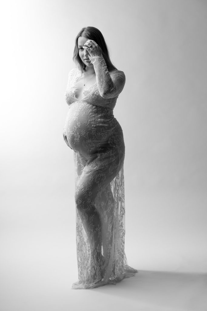 A pregnant woman stands gracefully in a bright studio setting for her maternity photos inspired by Vogue. She poses with one hand raised near her forehead and the other resting gently on her rounded belly while gazing directly toward the camera. Dramatic lighting creates strong highlights and shadows across the black and white image emphasizing the intricate lace patterns of her long fitted dress. The sheer fabric drapes elegantly around her form. She remains barefoot against the seamless white backdrop.