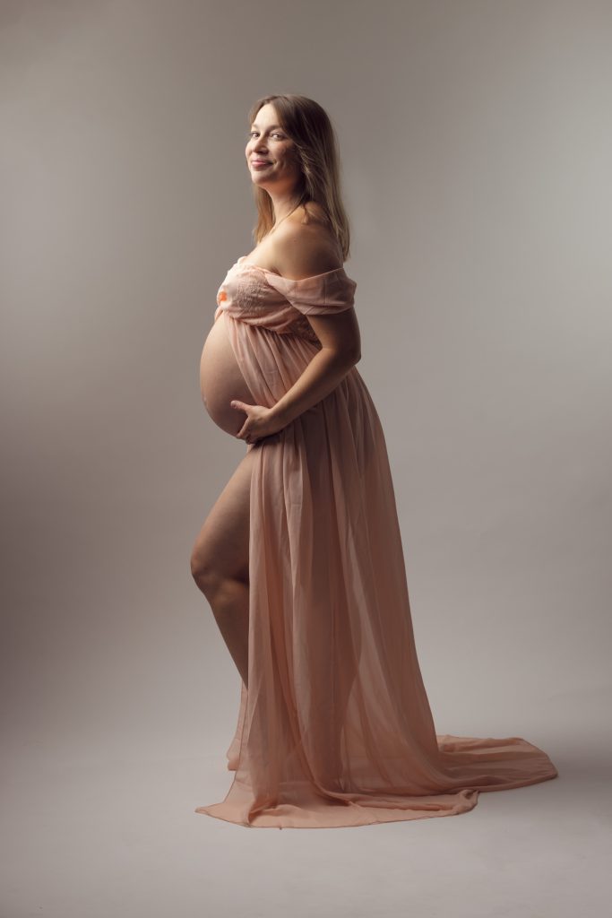 A pregnant woman stands gracefully in a softly lit studio setting for her maternity photos with open front dress. She turns slightly toward the camera with a warm smile while cradling her rounded belly with one hand. The flowing peach fabric drapes elegantly around her form with the open front creating soft movement and texture. Soft even lighting highlights the delicate tones of the dress and her natural silhouette. She remains barefoot against the minimalist backdrop.