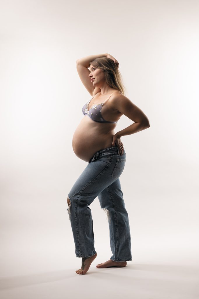 A pregnant woman stands in a bright studio setting during her maternity shoot in jeans. She poses in profile with one hand resting on her hip and the other raised gracefully to her hair while gazing to the side. Soft even lighting illuminates the natural colors of her light purple floral lace bra and blue ripped denim jeans. She remains barefoot against the seamless white backdrop.