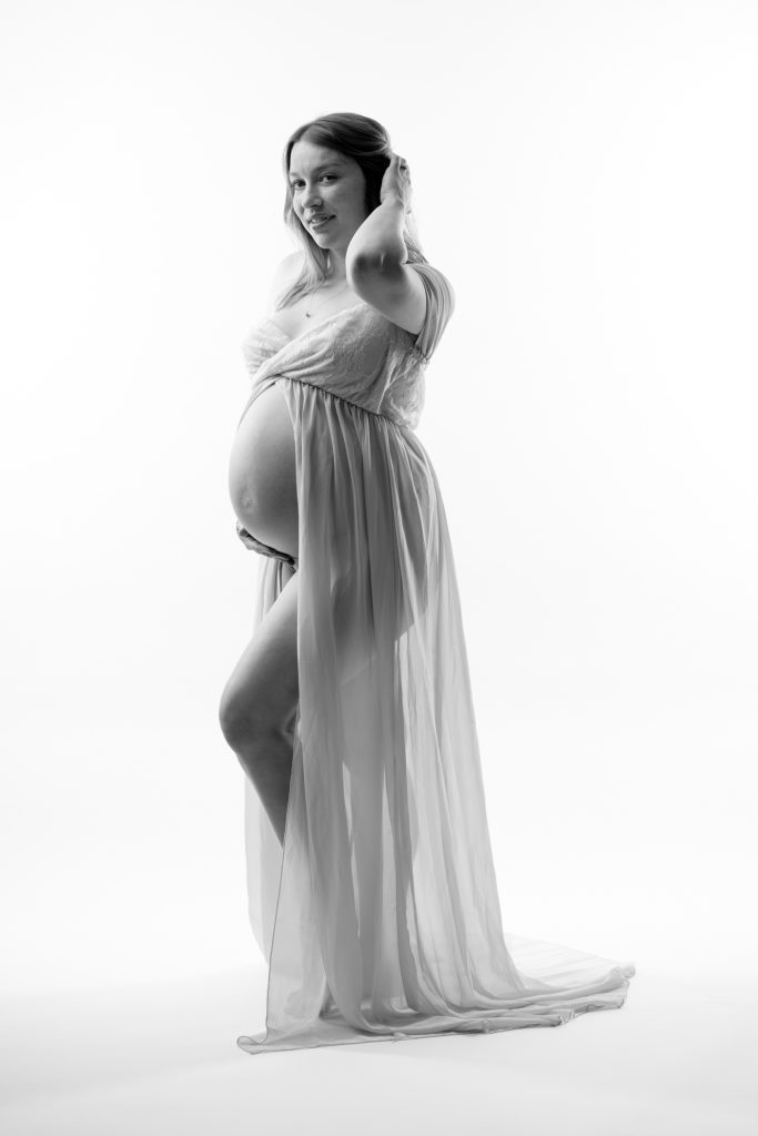A pregnant woman poses gracefully in a bright studio setting for her open front dress maternity photos. She stands with one hand raised to her hair while smiling toward the camera and cradling her rounded belly with the other hand. Dramatic lighting creates soft highlights and shadows across the black and white image emphasizing the flowing sheer fabric of her dress. The open front design drapes elegantly around her form with the long train pooling on the floor. She remains barefoot against the seamless white backdrop.