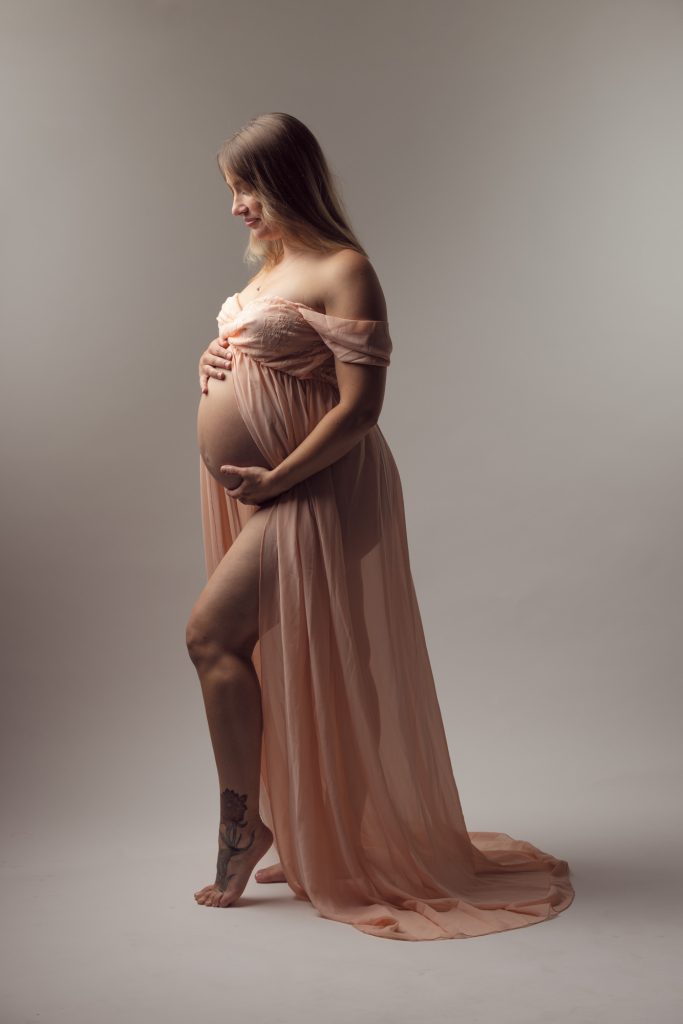 A pregnant woman stands in profile within a softly lit studio setting while wearing a pink open front maternity dress. She cradles her rounded belly gently with both hands and gazes downward in a serene pose. The flowing fabric drapes elegantly around her form creating beautiful movement in the image. Soft even lighting highlights the delicate peach tones of the dress and her natural silhouette. She remains barefoot with a visible tattoo on her ankle against the minimalist backdrop.
