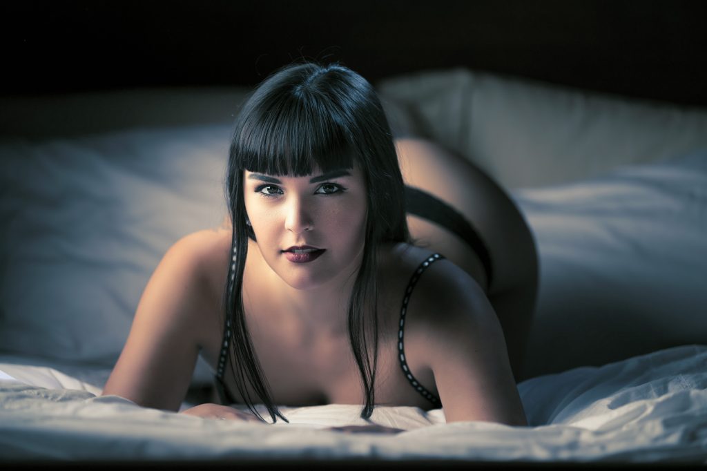 A woman with long dark hair and bangs lies on rumpled white bedding in a dimly lit bedroom demonstrating sexy boudoir photography poses. She props herself up on her elbows as she gazes directly at the camera wearing black lingerie. Cool dramatic lighting creates strong contrasts across the scene and adds depth to the composition.