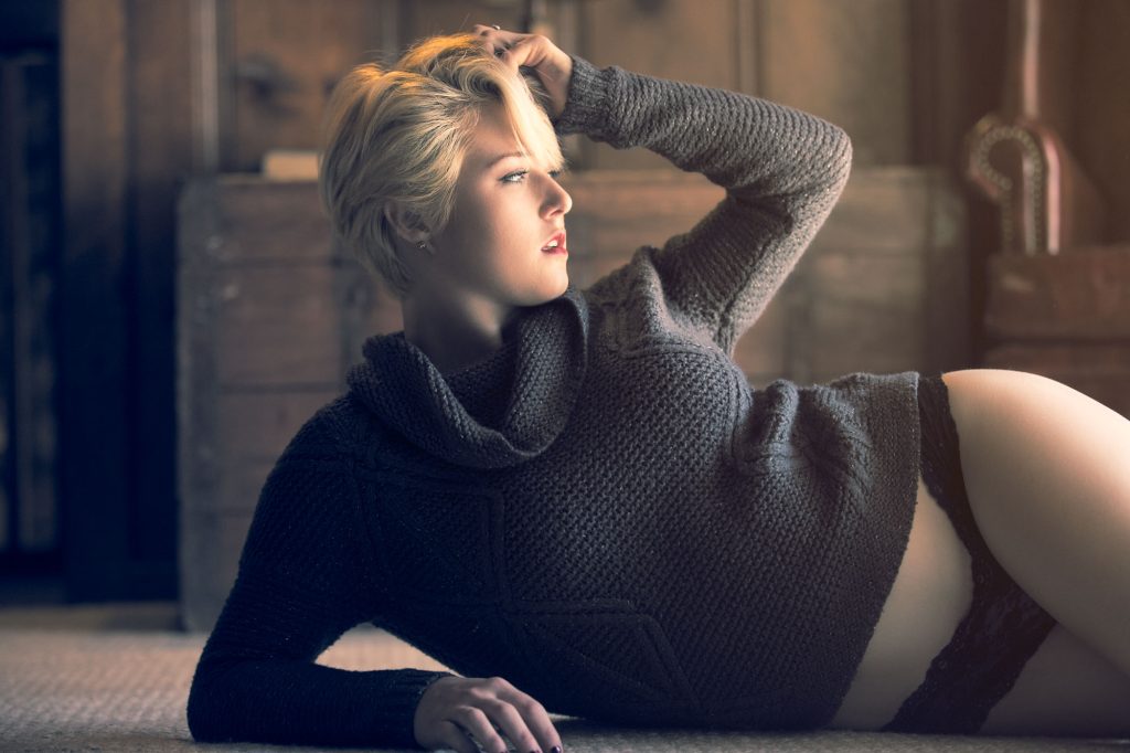A woman with short blonde hair showcases sexy poses for plus size women as she reclines on the floor in a warmly lit wooden interior. She props herself up on one arm with the other hand resting in her hair while gazing to the side and wearing a dark gray chunky knit turtleneck sweater paired with black lace underwear. Soft directional lighting highlights the textures of the knit fabric and creates gentle shadows across the rustic background in this intimate composition.